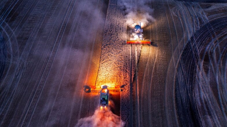 Mähdrescher und Traktorengespanne fahren nach Sonnenuntergang über ein Feld und dreschen Weizen. Die Ernte fällt schlechter aus als befürchtet.