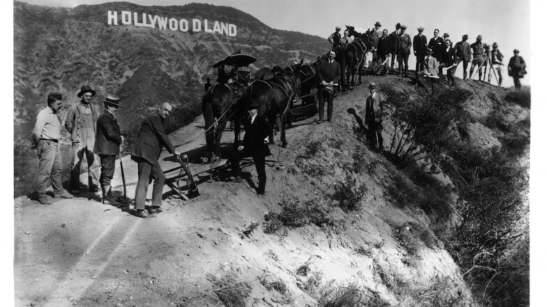 Menschen stehen vor dem übergroßen Schriftzug "HOLLYWOODLAND", der 1923 von einer Maklerfirma für Grundstücke als Werbegag in den damals unbewohnten Hügeln aufgestellt worden ist.