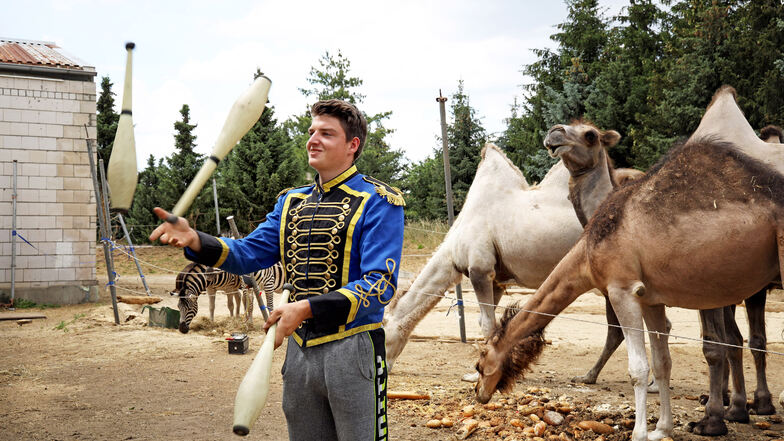 Adrian Schmidt jongliert in Paradeuniform vor der Futterstelle der Kamele und Dromedare. Dahinter befindet sich die Freiluft-Manege, wohin die Aeros-Zirkusleute zur öffentlichen Probe einladen.
