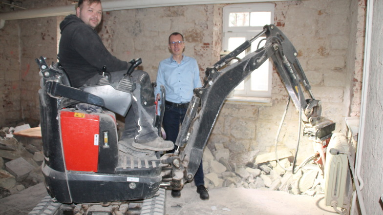 Bauarbeiter Oliver Lange bricht mithilfe eines Minibaggers und Stemmhammers den Steinfußbodenbelag auf. Bürgermeister Heiko Wersig (parteilos) macht sich ein Bild von den Arbeiten.