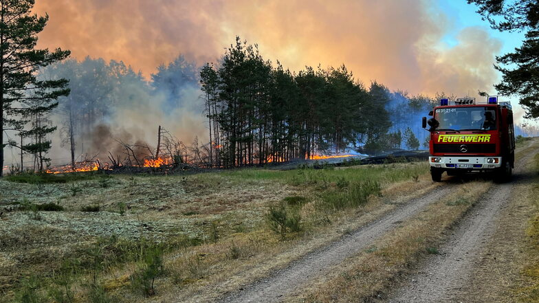 Rauch und Flammen steigen in den Himmel aus einem Waldgebiet bei Lübtheen, durch das ein Löschfahrzeug der Feuerwehr fährt.