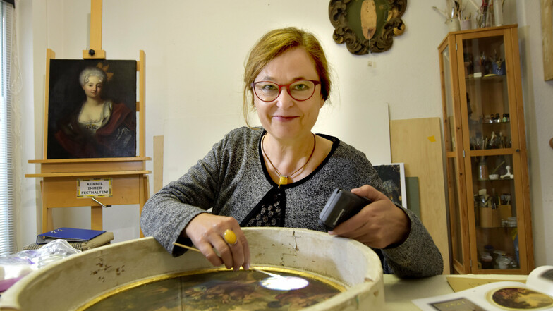 Die Restauratorin Anke Stenzel bei der Arbeit am Taufsteinbild in ihrem Atelier. Im Hintergrund ist das Gemälde aus der Rüstkammer mit Johanna Charlotte von Brandenburg-Schwedt zu sehen.
