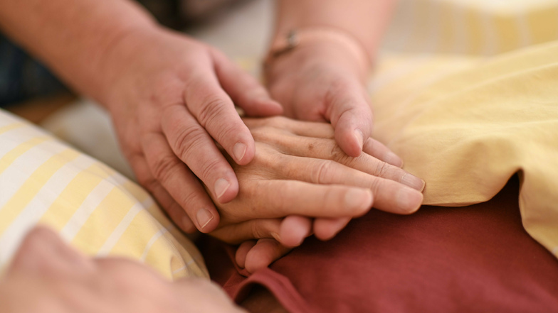Eine Hospizmitarbeiterin hält die Hand eines todkranken Menschen, der in einem Hospiz im Bett liegt. In Zeiten von Corona ist das leider nur schwer möglich.