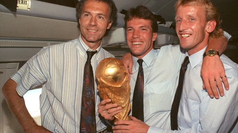 So sehen Weltmeister aus: Im Flugzeug beim Rückflug von Rom präsentieren der damalige DFB-Teamchef Franz Beckenbauer (l), Kapitän Lothar Matthäus (M) und Abwehrspieler Andreas Brehme den gewonnenen WM-Pokal.
