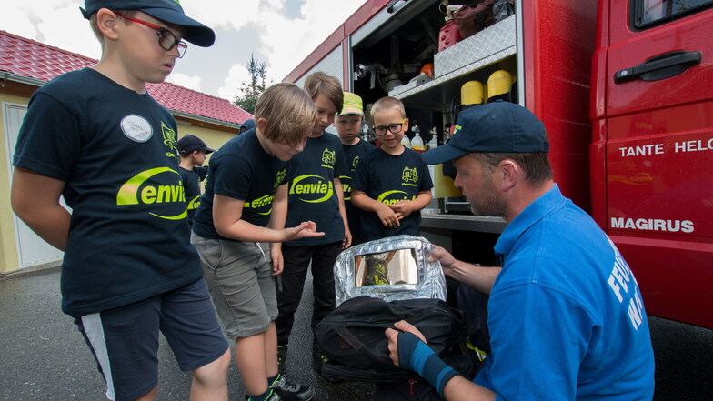 Stefan Voß, Ortswehrleiter von Meinsberg, zeigt den Mädchen und Jungen der Kinderfeuerwehr, welche besondere Ausrüstung bei starker Hitze neben den Atemschutzgeräten eingesetzt wird, wie der spezielle Flammschutz aus Aluminium.