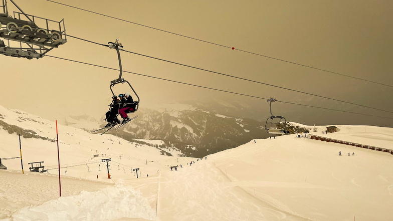 Skifahrer mit Gesichtsschutzmasken sitzen auf einem Sessellift, während Saharasand den Schnee und den Himmel orange färbt und eine besondere Lichtstimmung erzeugt.