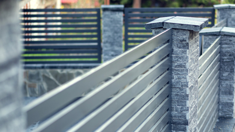 Geradlinig und modern umschließt dieser Zaun aus Metall gemeinsam mit den Steinsäulen das Grundstück auf einem Mauersockel.