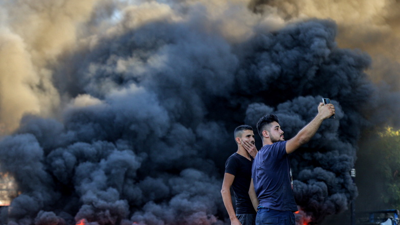 Libanon, Beirut: Zwei Männer machen ein Selfie vor brennenden Reifen, die eine Hauptverkehrsstraße blockieren, während eines Protestes gegen den massiven Anstieg der Benzinpreise. Die Kraftstoffpreise sind im Libanon inmitten einer akuten Finanzkrise in die Höhe gestiegen.