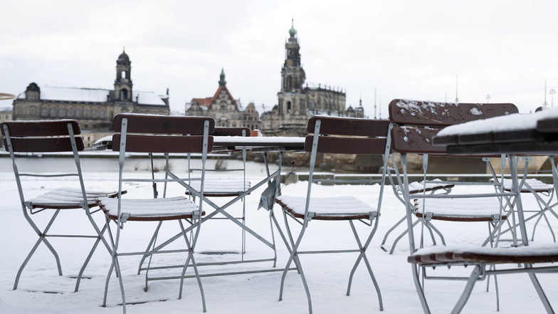 Mit Schnee bedeckt sind Tische und Stühle in einem Biergarten an der Elbe vor der Kulisse der Altstadt.