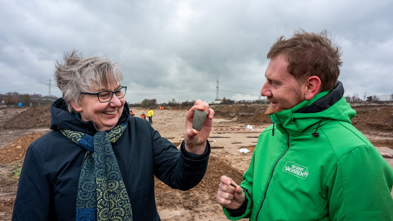 Regina Smolnik, Chefin des Landesamtes für Archäologie, zeigt Ministerpräsidenten Michael Kretschmer (CDU) ein Steinbeil, das auf dem Grabungsareal von Karls Erlebnis-Dorf gefunden wurde.