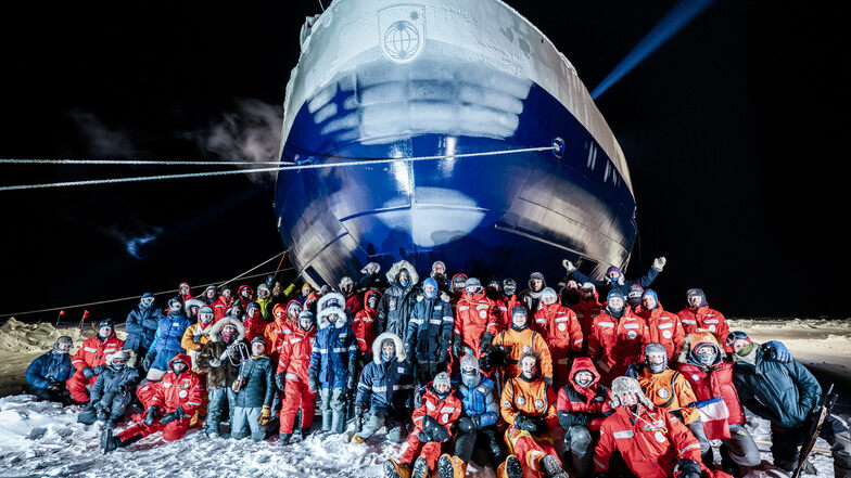 Die derzeitige Mannschaft der Mosaic Forschungsgruppe auf der Polarstern hat sich zu einem Gruppenfoto vor dem Bug vom eingefrorenen Forschungsschiff aufgestellt.