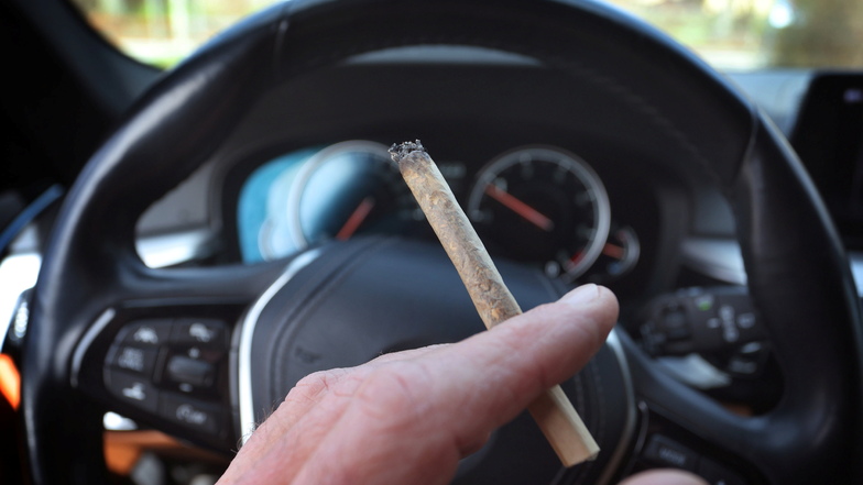 Ein Mann sitzt mit einem Joint zwischen den Fingern am Steuer eines Autos.