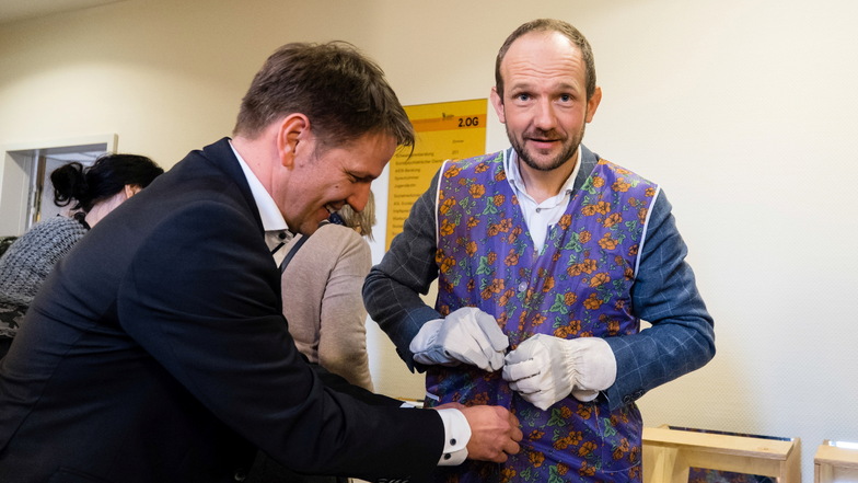 Staatssekretär Sebastian Vogel (links) hilft Landrat Stephan Meyer eine Schürze anzuziehen. Das Experiment mit den groben Handschuhen simuliert schlechte Feinmechanik im Alter und war am Dienstag im Kreis-Gesundheitsamt auszuprobieren.