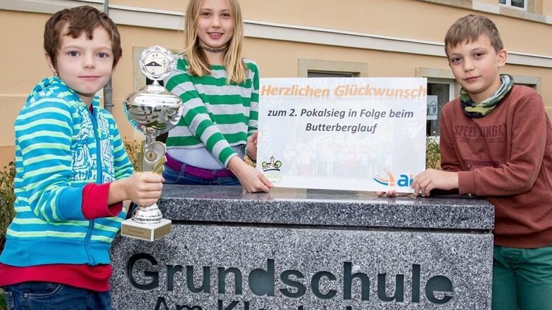 Jamie Suchan, Luise Wiltgrupp und Michael Symmank (von links) zeigen den Pokal und das Schild für den zweiten Sieg in Folge beim Butterberglauf der Grundschulen.