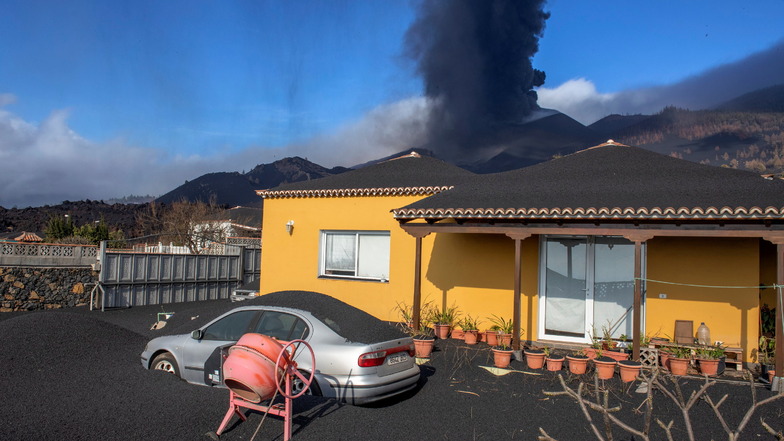 Haus und Wagen stehen unter Asche, während der Vulkan im Hintergrund weiter Aschenwolken und Lava ausstösst.