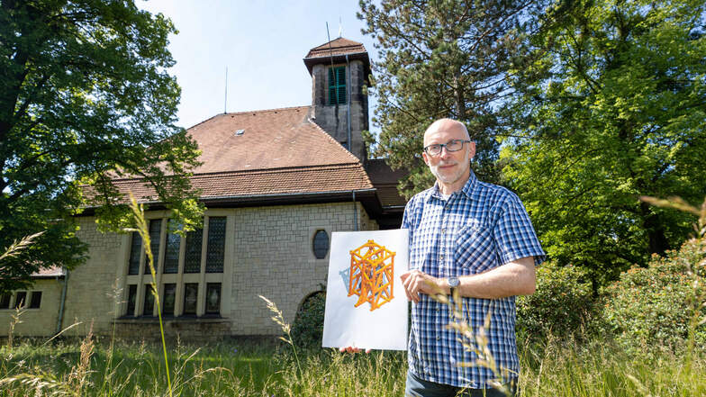Pfarrer Burkhard Nitzsche steht auf einer Wiese mit der Graupaer Kirche im Hintergrund. In der Hand hat er eine Zeichnung des neuen Glockenstuhls.