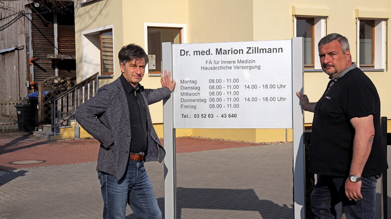 Der Wülknitzer Bürgermeister Hannes Clauß und CDU-Gemeinderat Sven Bauer stehen vor der Tafel der Arztpraxis von Dr. Marion Zillmann. Sie ist geschlossen.