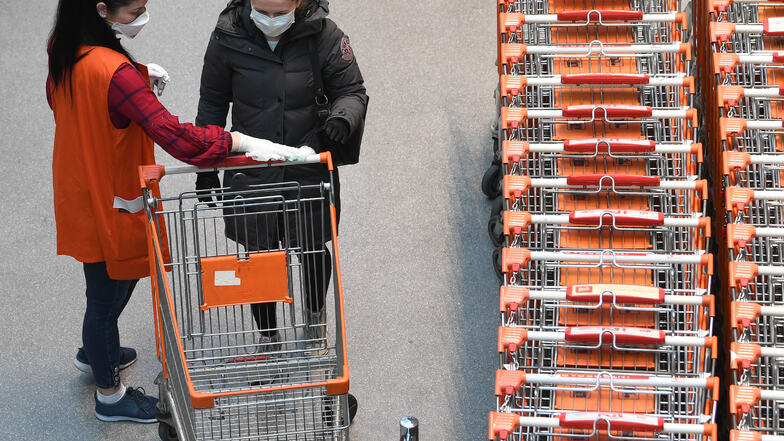 Die Mitarbeiterin eines Interspars desinfiziert den Einkaufswagen einer Kundin. In den meisten Lebensmittelgeschäften und Drogerien in Österreich müssen die Menschen aufgrund der Coronavirus-Krise nun einen Mundschutz tragen. 