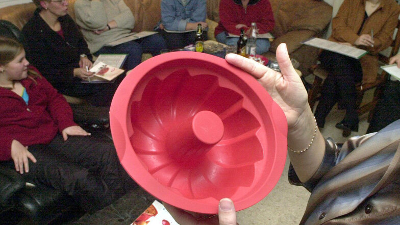 Eine Tupperware-Beraterin preist eine Kunststoffbackform einer Runde von interessierten Kundinnen in einem Wohnzimmer an.