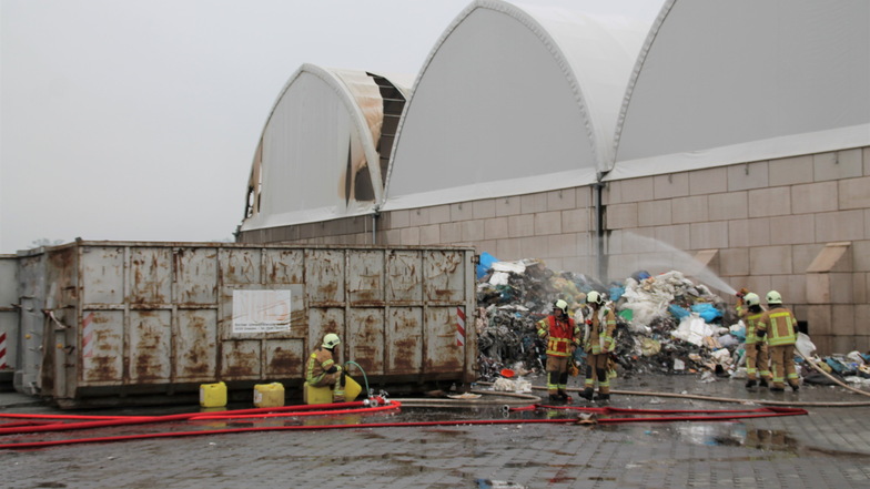 Am Mittwochvormittag haben die alarmierten Feuerwehrleute den ins Freie beförderten Müll weiter bewässert. Im Hintergrund: die beschädigte Lagerhalle.