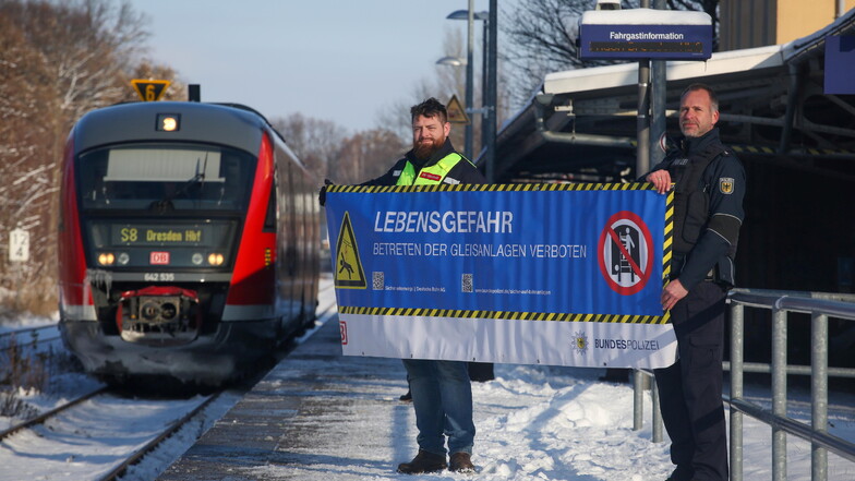Die Präventionsbeauftragten von Bahn und Bundespolizei, Bastian Peter (l.) und Patrick Seiler, machen am Pulsnitzer Bahnhof mit einem Banner auf die Gefahr der illegalen Gleisübertritte aufmerksam.