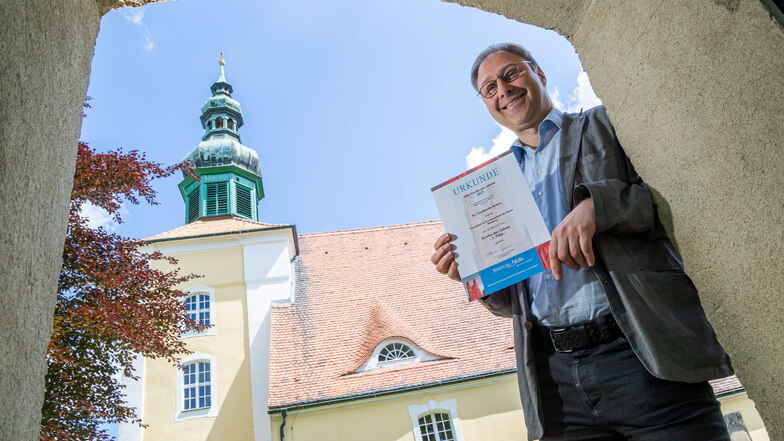 Pfarrer Daniel Jordanov steht vor der evangelischen Kirche in Klitten und zeigt die Urkunde für den dritten Platz im Wettbewerb der KiBa-Stiftung.