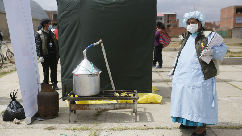 Bolivien, El Alto: Eine Mitarbeiterin des Ministeriums für Traditionelle Medizin steht in Schutzanzug neben einer Dampfkabine, die während der Corona-Pandemie zur Vorbeugung von Erkrankungen der Atemwege an einer Ecke aufgestellt wurde.