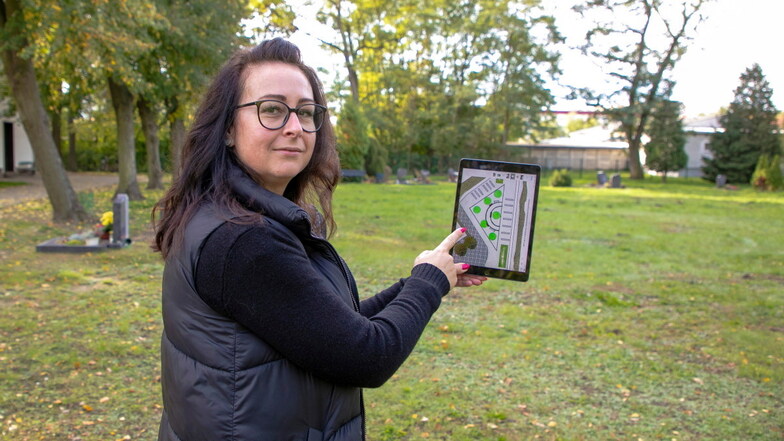 Hauptamtsleiterin Tina Noack zeigt auf dem Tablet, wie der neue Urnenwald auf dem Gröditzer Friedhof Wainsdorfer Straße geplant ist. Die sechs Punkte sind die Bäume, die im Herbst gepflanzt werden sollen.