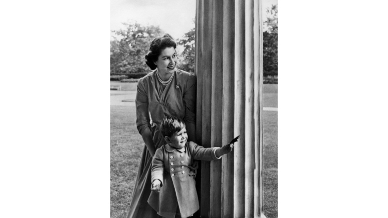 1950 schaut Königin Elizabeth II. mit dem zweijährigen Sohn, Prinz Charles, hinter einer Säule, auf dem Grundstück vom Clarence House, hervor.