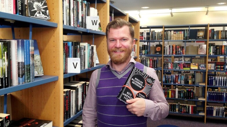 Mladen Vukovic mit einem Buch, das er gerade privat liest, an einem Regal der Brigitte-Reimann-Stadtbibliothek Hoyerswerda. Es ist nicht ausgeschlossen, dass unter „V“ eines Tages ein Science-Fiction-Roman aus der Feder von Mladen Vukovic stehen wird.