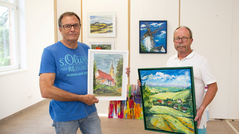 Jörg Arnhold (links) und Gunter Hauswald, hier mit Ansichten von Possendorf, bereiten eine Ausstellung von Hobbykünstlern in der Pfarrscheune des Bannewitzer Ortsteiles vor. Im Hintergrund ist eine der Galeriewände für die über 100 Arbeiten zu sehen.