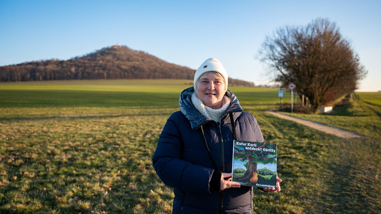 Kerstin Poost steht mit dem von ihr geschriebenen Kinderbuch „Kater Karli entdeckt Görlitz“ beim Trimm-Dich-Pfad in Schlauroth vor der Landeskrone. In dem Buch geht es um den Görlitzer Hausberg.