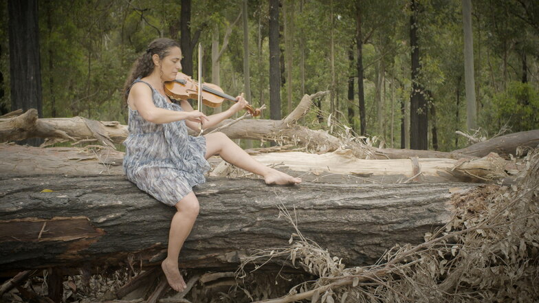 Bianca aus Australien spielt Geige auf einem verbrannten Baumstamm, der nach einem verheerenden Feuer zurückgeblieben ist.