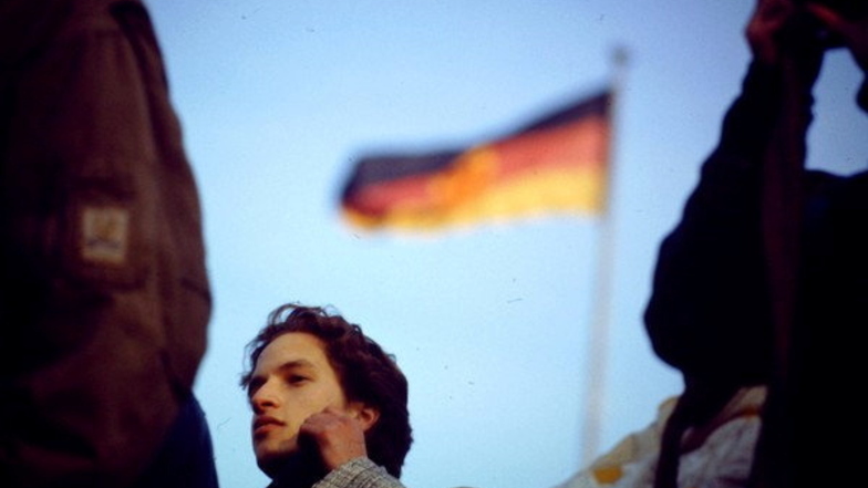 Ein junger Mann, offensichtlich fassungslos von dem Mauerfall, steht vor einer DDR-Fahne in Berlin. Auch 33 Jahren später beschäftigt die Wiedervereinigung die Teilnehmer beim Bautzen-Forum.