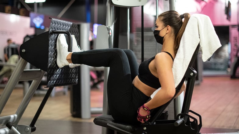 Eine Frau trainiert mit Mundschutz in einem Fitnessstudio an einer Beinpresse.