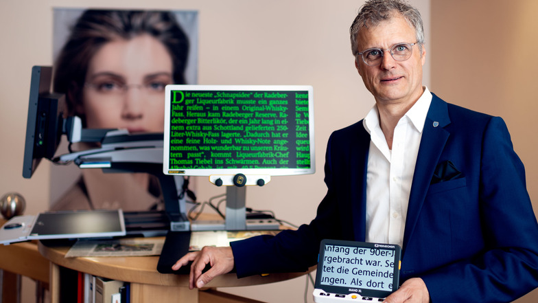 Augenoptikermeister Michael Plüschke zeigt eine elektronische Lupe. Menschen mit Sehbehinderung können sie jederzeit mitführen. Im Hintergrund ist ein Bildschirmlesegerät zu sehen.