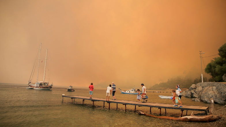 Türkei, Bodrum: Touristen stehen während eines Waldbrands auf einem Steg und warten darauf, evakuiert zu werden.
