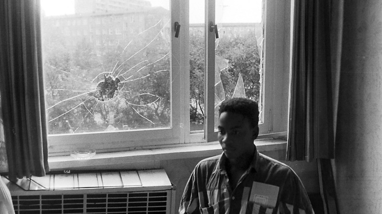 Ein mosambikanischer Vertragsarbeiter in seinem Wohnheimzimmer, im Hintergrund die eingeworfenen Fensterscheiben.