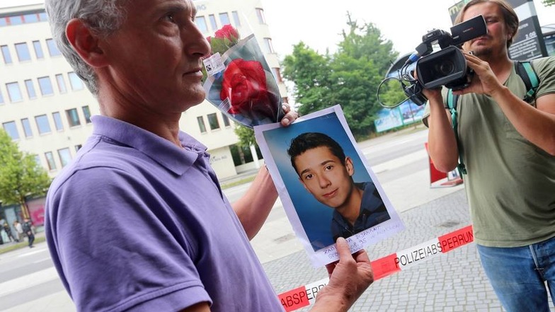Naim Zabergja steht mit einem Foto seines getöteten Sohnes Dijamantvor Medienvertretern am Olympia-Einkaufszentrum in München (Bayern), das die Polizei nach einer Schießerei mit Toten und Verletzten am Vortag abgesperrt hat.