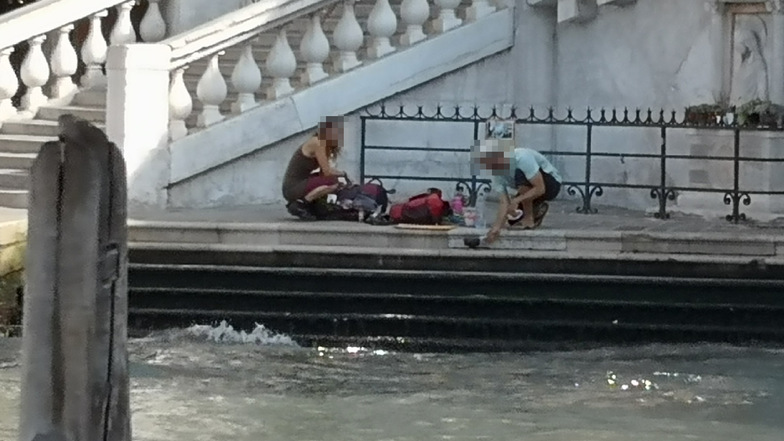 Zwei deutsche Touristen sitzen mit ihrem Gepäck an der Treppe der Rialto-Brücke und kochen sich mit einem kleinen Kaffeekocher einen Kaffee auf dem Gehweg.
