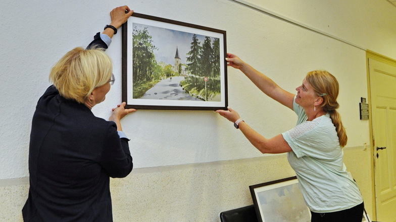 Bürgermeisterin Anita Maaß (links) und Susann Kohlmann von der Stadtverwaltung hängen eines der Bilder im Rathaus auf. Auf diesem ist die Kirche in Striegnitz zu sehen.