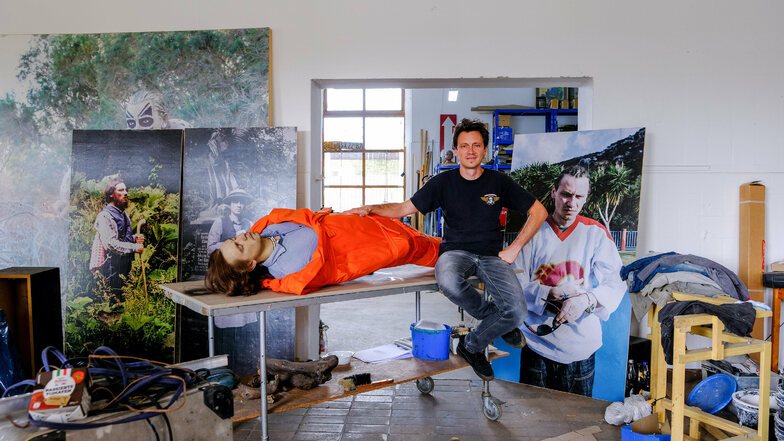 Umgeben von mehreren seiner Arbeiten wie plakatgroßen Fotografien sowie einer Leichenskulptur, dessen Gesicht die Züge des Künstlers trägt, sitzt Manuel Frolik auf einem Rolltisch im großen Atelierraum.