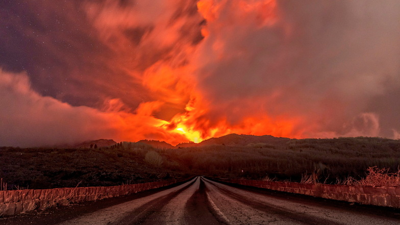 Ein glühender Lavastrom fließt über den Ätna, der von Rauch umgeben und über dem ein rötlich gefärbter Himmel zu sehen ist.