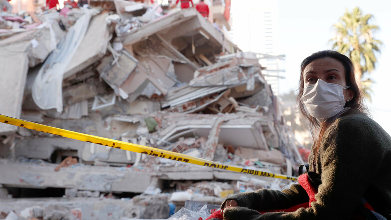 31.10.2020, Türkei, Izmir: Eine Anwohnerin, die sich aus Angst vor Nachbeben im Freien aufhält, beobachtet Rettungskräfte in den Trümmern eines eingestürzten Gebäudes nach Überlebenden suchen.