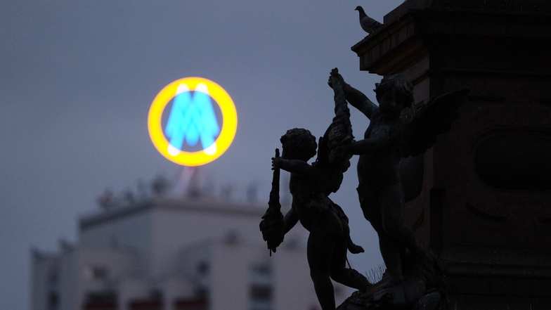 Das Logo der Leipziger Messe leuchtet an einem Morgen auf dem Wintergartenhochhaus, während im Vordergrund eine Figur des Mendebrunnens sowie eine rastende Taube zu sehen sind.