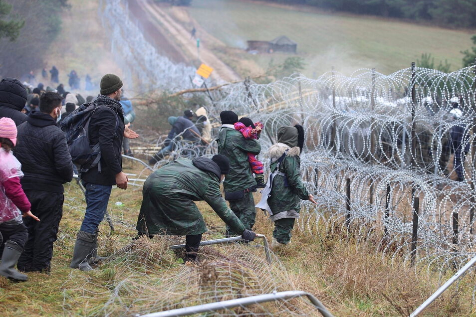 Imigranci burzą ogrodzenie na granicy białorusko-polskiej, a policja stoi za ogrodzeniem z drutu kolczastego.